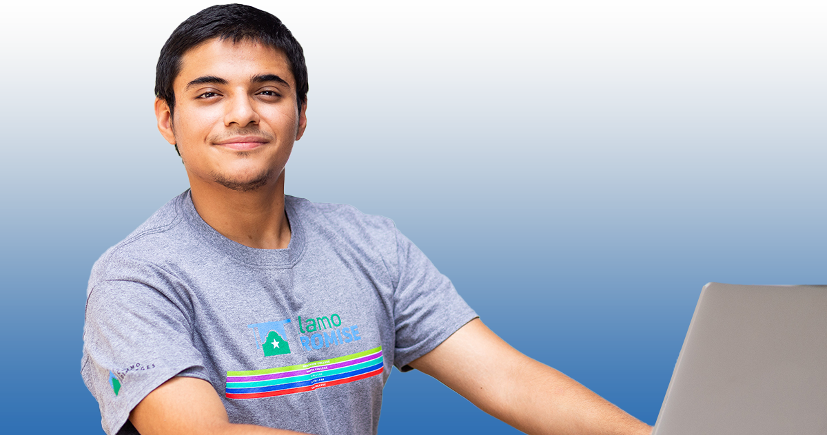 Male student smiling while working on a laptop; student is wearing a grey Alamo Promise shirt with the logo on the front